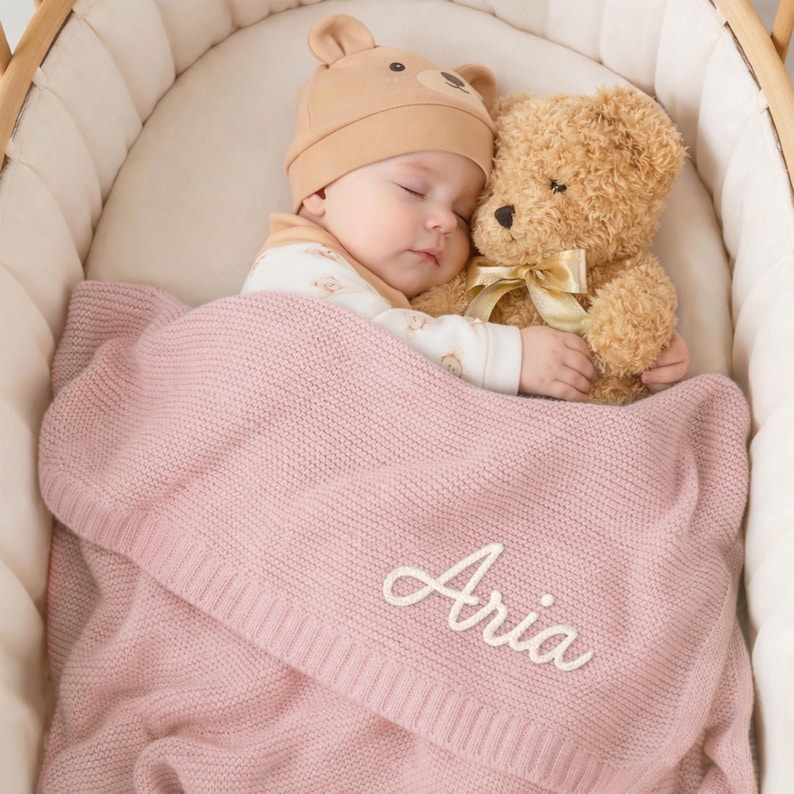 May include: A sleeping baby wearing a tan bear hat and a white onesie, nestled in a crib with a teddy bear. A pink knitted blanket with the name "Aria" embroidered in white covers the baby.