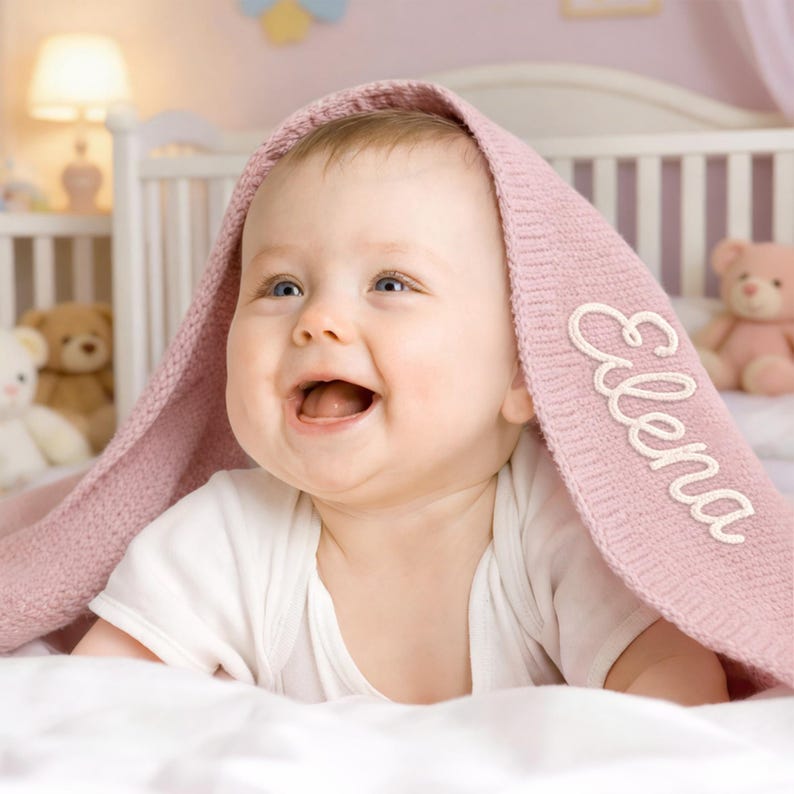 May include: A smiling baby is wrapped in a soft, pink blanket with the name "Elena" embroidered in white. The baby is wearing a white onesie and is lying on a white surface. A teddy bear and a lamp are visible in the background.