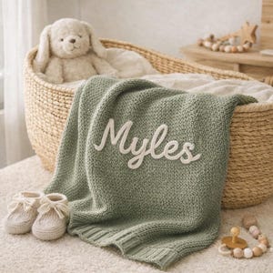 May include: A sage green knitted baby blanket with the name "Myles" embroidered in white. The blanket is draped over a woven basket, with a stuffed animal and baby shoes nearby. A wooden teething toy is also visible.