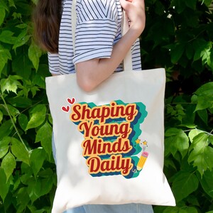 May include: A white tote bag with a retro-style design featuring the text "Shaping Young Minds Daily" in red and yellow lettering. The design also includes a pencil and stars.