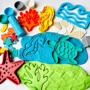 May include: A collection of colorful play dough tools and molds. Includes a blue roller, various sea creature molds like a seahorse, turtle, and starfish, and wave-shaped cutters. The play dough is blue and green, with molded sea creatures.
