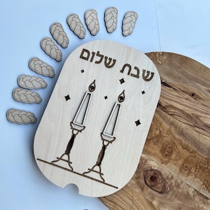 May include: A wooden board with the Hebrew words "Shabbat Shalom" and a design of two candlesticks with candles. The board is surrounded by eight small wooden challah bread shapes.