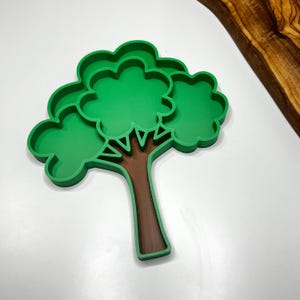 May include: A green tree-shaped cookie cutter with a brown trunk. The cookie cutter has a detailed design with a rounded top and a sturdy base. The cutter is made of plastic and is designed for baking cookies or other treats.