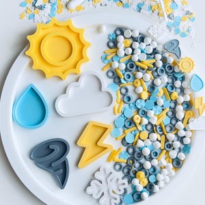 May include: A white plate holds weather-themed cookie cutters and sprinkles. The cutters include a yellow sun, blue raindrop, white cloud, yellow lightning bolt, gray tornado, and white snowflake. Sprinkles are blue, yellow, white, and gray.