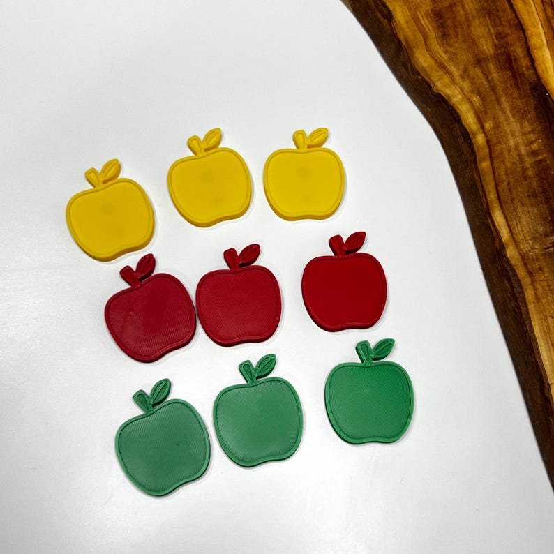 May include: Nine apple-shaped cookie cutters in yellow, red, and green. Each cutter has a leaf and stem detail. The cutters are arranged on a white surface with a wooden board in the background. Great for baking.