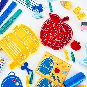 May include: A collection of colorful school-themed items, including a yellow school bus, blue backpacks, red apples, and crayons. There are also small paper airplanes, scissors, and number blocks. The image has the text "WHAT'S IN MY BACKPACK?"