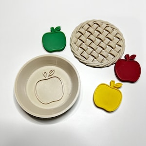 May include: An assortment of apple-themed baking tools. Includes a beige pie crust topper, a beige pie dish with an apple outline, and green, red, and yellow apple-shaped cookie cutters. Perfect for creating apple pies and treats.