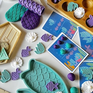 May include: A selection of mermaid-themed craft materials. Includes teal, purple, and white molds, a wooden tray, and colourful play dough. Also features cards with mermaid and fish illustrations, ideal for creative projects.