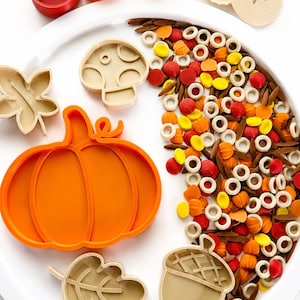 May include: A white circular tray containing a large orange pumpkin-shaped cutter, beige leaf, mushroom, and acorn cutters, and small beige, red, orange, and yellow autumn-themed sensory filler pieces, alongside several colorful plastic cups.