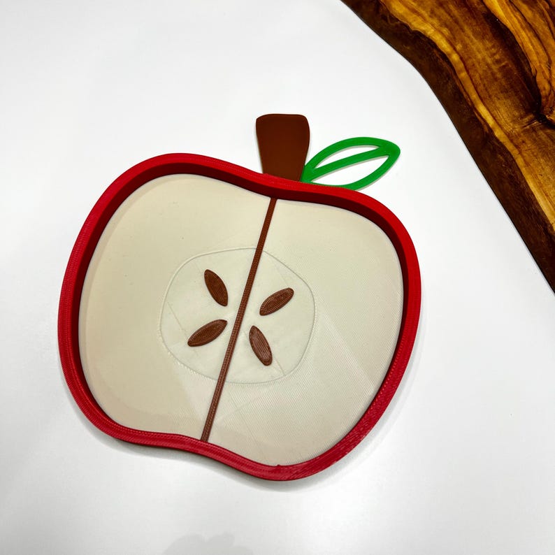 May include: A red, white, and brown apple-shaped object. The object is a cross-section of an apple, with a red outline, a white interior, and brown seeds. A brown stem and green leaf are attached to the top. The object is on a white surface.