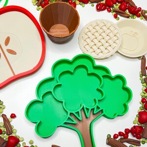May include: A collection of colorful, food-themed cookie cutters and decorative elements. Includes red, green, and yellow apple shapes, a tree, a pie crust, a pie plate, a wooden barrel, and decorative elements.