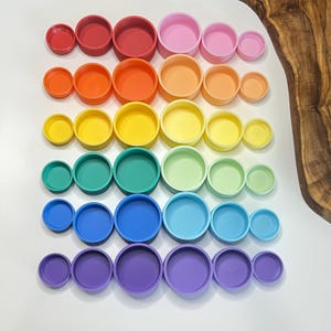 May include: A collection of colorful, round containers arranged in rows by color. The containers range in size and include shades of red, orange, yellow, green, blue, and purple. The containers are made of a solid material and have a simple, modern design.
