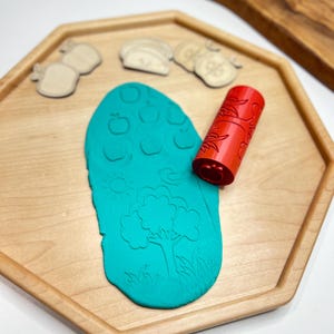 May include: A wooden tray holds a teal-colored playdough with embossed apple and tree designs. A red roller with a floral pattern and wooden fruit-shaped stamps are also present. The tray is light-colored wood.