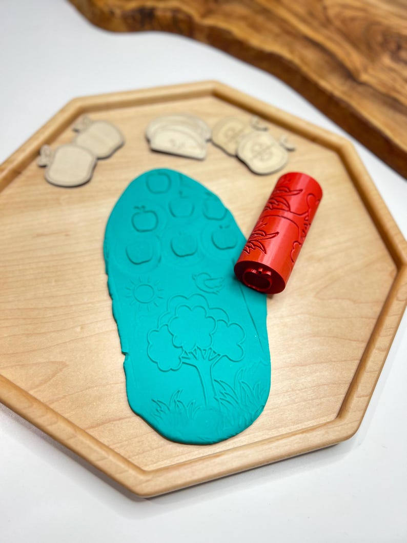 May include: A wooden tray holds a teal-colored playdough with embossed apple and tree designs. A red roller with a floral pattern and wooden fruit-shaped stamps are also present. The tray is light-colored wood.