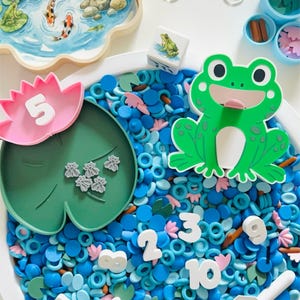 May include: A sensory play set with a pond theme. Includes a wooden tray with a pond scene, a lily pad with the number 5, a green frog, blue and pink water beads, and number tokens. Educational toy.