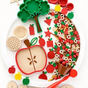 May include: A sensory play set featuring a large white circular tray containing a green tree-shaped cutter, a red apple-shaped plate, small red bowls, a brown bucket, a red scoop, and various wooden and plastic shapes including apples, stars, and trees.
