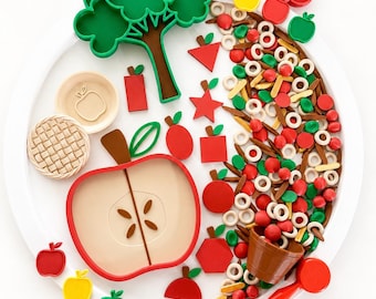 May include: A sensory play set featuring a large white circular tray containing a green tree-shaped cutter, a red apple-shaped plate, small red bowls, a brown bucket, a red scoop, and various wooden and plastic shapes including apples, stars, and trees.