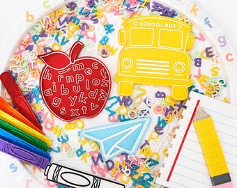 May include: A circular white tray filled with colorful small plastic letters, featuring a large red apple shape with letters, a yellow school bus, a blue paper airplane, a pencil-shaped notepad, and a black and white marker with crayons.