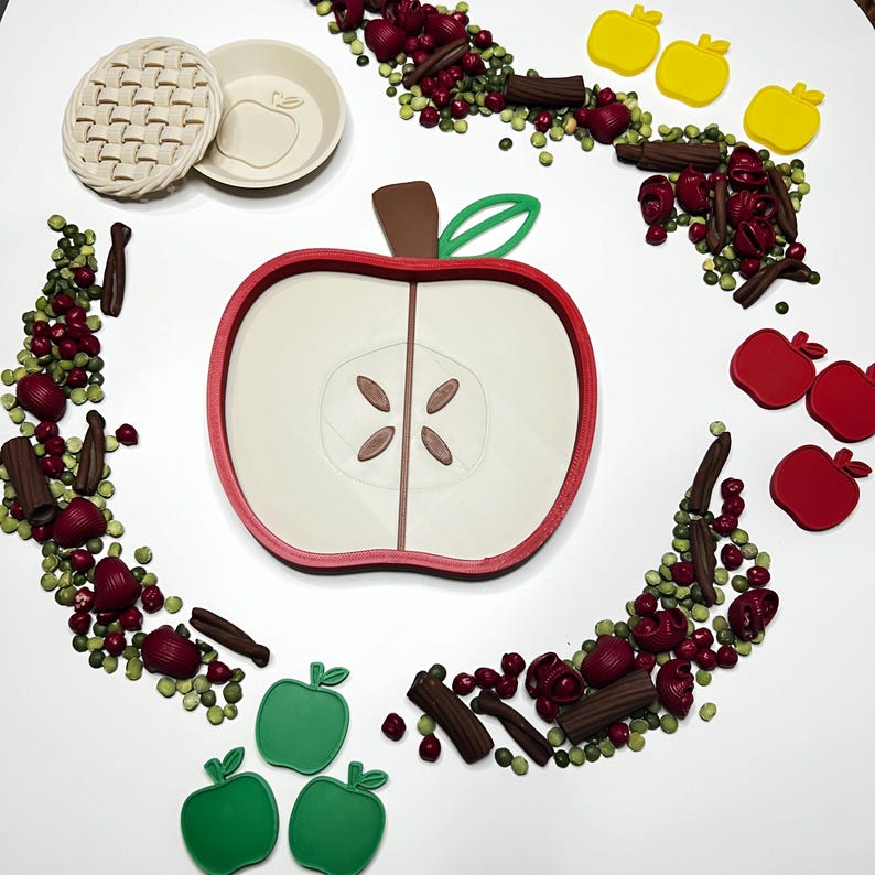 May include: An overhead shot features a large apple-shaped mold with a red outline, alongside various apple-themed cookie cutters in red, green, and yellow. Decorative elements include cinnamon sticks and red candies, creating a festive, food-related theme.