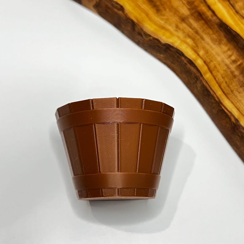 May include: A small, brown, barrel-shaped container with vertical lines and a slightly flared shape. The container is made of a solid material and has a smooth surface. The background is white with a wooden cutting board.