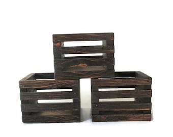 May include: Three dark brown wooden crates stacked on top of each other. The crates have a rustic look and are made of unfinished wood.