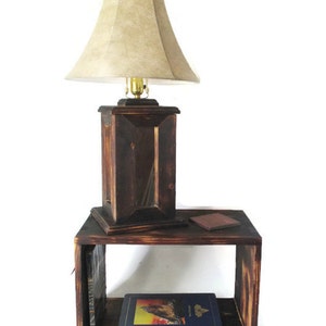 May include: A dark brown wooden lamp with a beige lampshade sits on a dark brown wooden end table with two shelves. A book is on the bottom shelf and a book and a small brown object are on the top shelf.