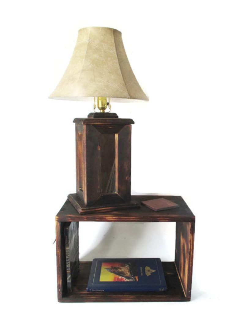 May include: A dark brown wooden lamp with a beige lampshade sits on a dark brown wooden end table with two shelves. A book is on the bottom shelf and a book and a small brown object are on the top shelf.