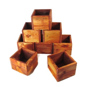 May include: A set of eight wooden boxes stacked in a pyramid shape. The boxes are made of natural wood with a reddish-brown stain. The boxes are all the same size and shape, and they have a rustic look.