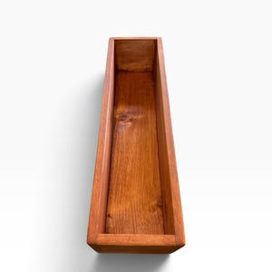 May include: A long, rectangular wooden planter box with a warm, medium-brown stained finish. The container features visible wood grain patterns and natural knots on the interior base, with straight, clean edges and a hollow, open-top design.