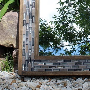 May include: A rectangular mirror with a wooden frame and a mosaic tile border. The tiles are in shades of gray and brown. The mirror is reflecting a view of trees and a blue sky.