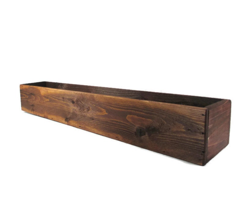 Puede incluir: Una jardinera de madera rectangular y larga, con una mancha de color marr&oacute;n oscuro. La jardinera est&aacute; hecha de madera natural con grano y nudos visibles.