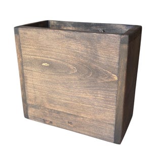 May include: A rectangular wooden box with a dark brown stained finish. The box features visible wood grain patterns on the front panel and sturdy vertical corner joints. The top is open, revealing the hollow interior of the container.