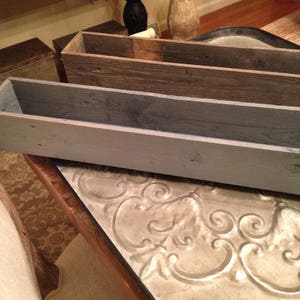 Puede incluir: Dos jardineras de madera, una gris y otra marr&oacute;n, est&aacute;n apiladas una encima de la otra. Las jardineras est&aacute;n hechas de madera reciclada y tienen un aspecto r&uacute;stico. Las jardineras est&aacute;n sobre una mesa con una superficie de metal decorativa.