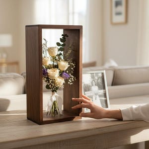 May include: A wooden display case with a bouquet of white and purple flowers. The flowers are arranged in a clear vase and tied with a white ribbon. The case is rectangular and sits on a light-colored wooden table. A framed photo is next to the case.