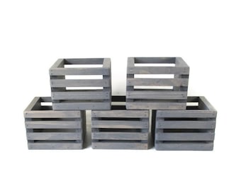 May include: Five gray wooden crates arranged in a cluster. Each crate is square with horizontal slats and a sturdy frame. The crates are painted in a muted gray color, offering a rustic and versatile aesthetic. Ideal for storage or display.