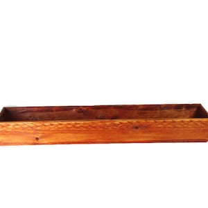 May include: A long, rectangular wooden planter box with a natural wood finish. The planter is made of unfinished wood and has a rustic look.