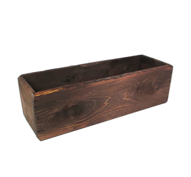 May include: A dark brown wooden planter box with a rectangular shape. The planter is made of unfinished wood and has a rustic look.
