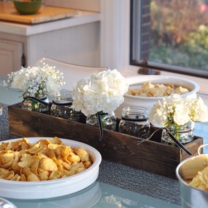 Puede incluir: Un macetero de madera r&uacute;stico con tres tarros de vidrio llenos de flores blancas. Los tarros tienen una cinta negra atada alrededor. El macetero est&aacute; sentado en una mesa con un taz&oacute;n blanco de patatas fritas y un taz&oacute;n blanco con una sustancia marr&oacute;n.