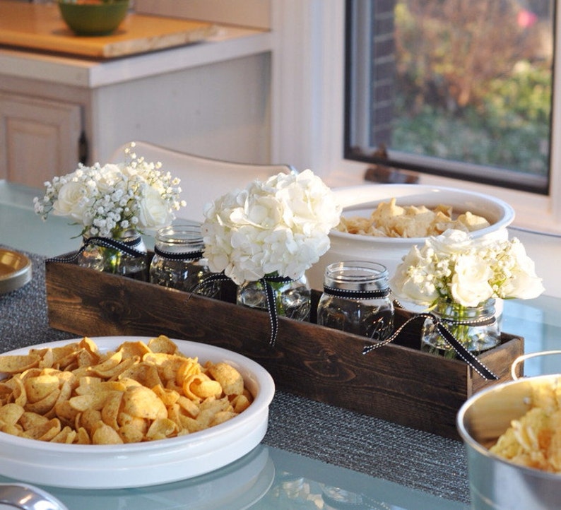 Puede incluir: Un macetero de madera r&uacute;stico con tres tarros de vidrio llenos de flores blancas. Los tarros tienen una cinta negra atada alrededor. El macetero est&aacute; sentado en una mesa con un taz&oacute;n blanco de patatas fritas y un taz&oacute;n blanco con una sustancia marr&oacute;n.
