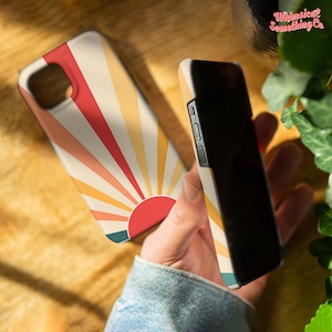 May include: Two phone cases, one with a retro sunburst design in red, orange, and yellow, and the other black. The sunburst case has a red sun in the centre.