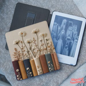 May include: A Kindle e-reader case featuring a beige cover with embroidered white daisy-like flowers and a row of vertical fabric book spines in shades of burgundy, mustard, navy, tan, brown, and sage green, each with small floral embroidery.