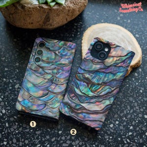 May include: Two phone cases with a iridescent abalone shell pattern. The cases are both black and have a camera cutout. The case on the left has three camera holes and the case on the right has two camera holes.