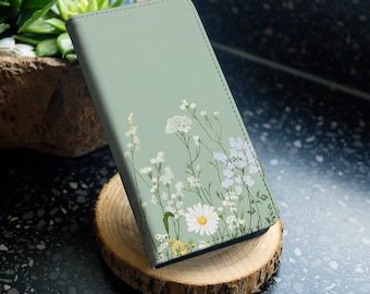 Könnte beinhalten: Eine salbeigrüne Handyhülle mit einem Blumendesign mit weißen Blüten und grünen Blättern. Die Hülle hat eine dezente Textur und eine matte Oberfläche.