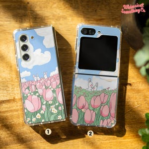 May include: Two clear phone cases with a whimsical design featuring pink tulips, green grass, and white bunnies against a blue sky. The case on the left is for a standard phone, while the case on the right is for a flip phone.
