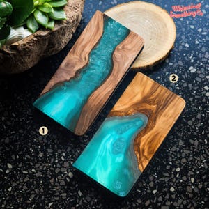 Könnte beinhalten: Zwei Handyhüllen mit einem Design aus Holz und blauer Harz. Die Hüllen haben ein natürliches Holzmaserungsmuster mit einem blauen Harzfluss, der durch die Mitte fließt. Der Text "Whimsical Something Co." befindet sich in der oberen rechten Ecke des Bildes.