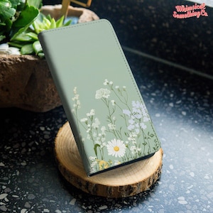 以下が含まれることがあります： セージグリーンの長方形のスマートフォンケース。白い野花とマーガレットが描かれた繊細なボタニカルプリントが特徴です。細い緑の茎と小さな白い花が表面全体に散りばめられており、Whimsical Something Co.というテキストが記されています。