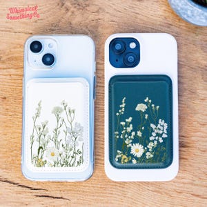 Könnte beinhalten: Zwei weiße Handyhüllen mit floralen Designs. Eine Hülle hat einen weißen Hintergrund mit einem floralen Design, und die andere hat einen dunkelblauen Hintergrund mit einem floralen Design. Die Hüllen liegen auf einer Holzoberfläche.