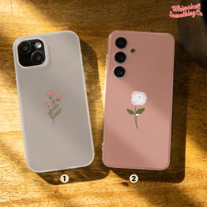 Könnte beinhalten: Zwei Handyhüllen mit Blumenmustern. Die Hülle links ist weiß mit einem rosa Blumendesign. Die Hülle rechts ist rosa mit einem weißen Blumendesign.