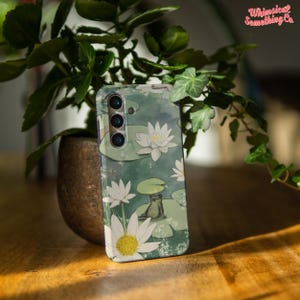 May include: A green, white, and brown phone case with a frog sitting on a lily pad and white flowers. The case has a camera lens cutout.
