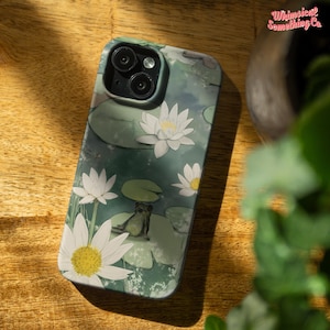 May include: A phone case with a green, blue and white watercolour design featuring lily pads, water lilies and a frog. The case has a camera cutout and a white background.